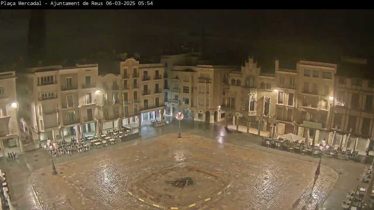 A rain-soaked town square at night is surrounded by multi-story buildings with outdoor seating areas partially occupied.