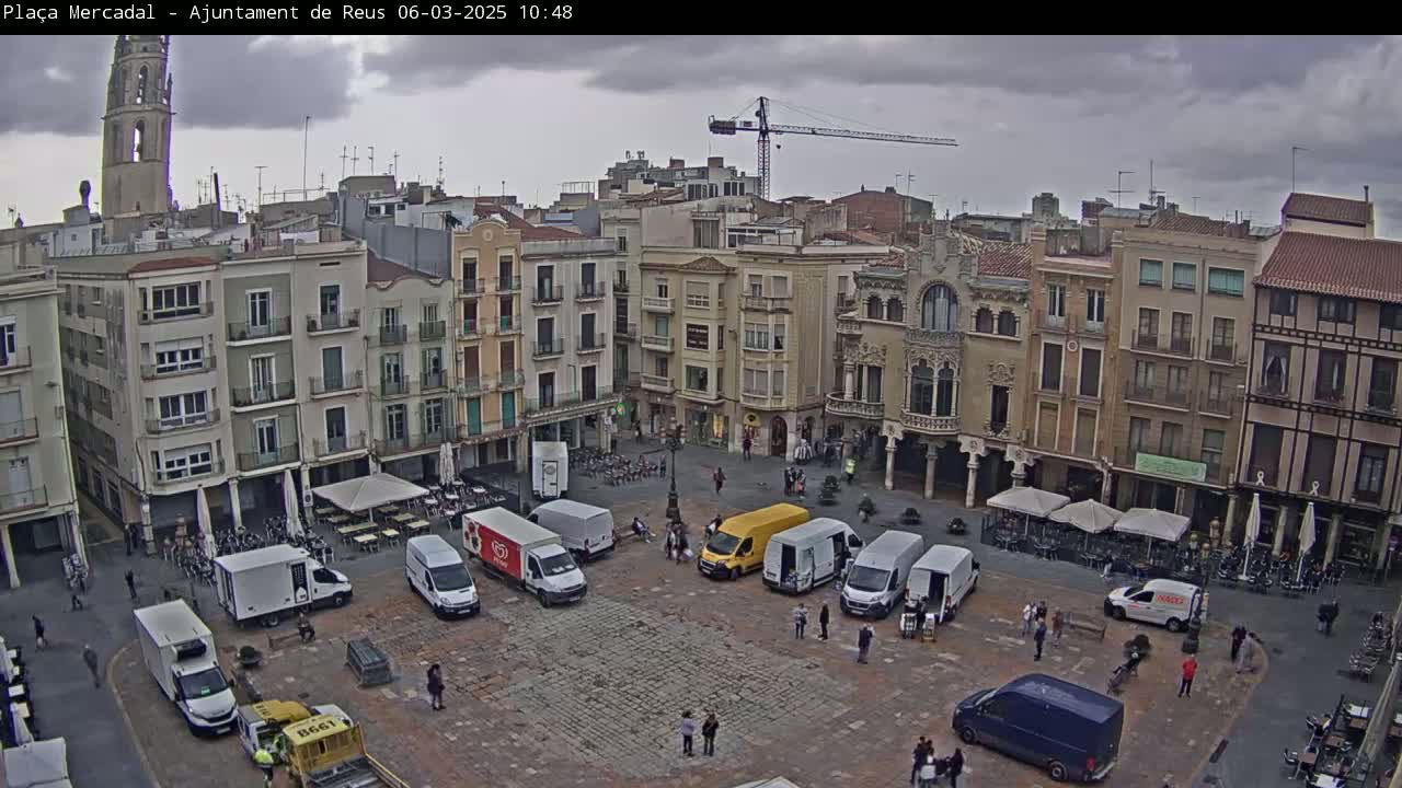 A town square, under an overcast sky, is filled with parked delivery vans and several people, surrounded by multi-story buildings.