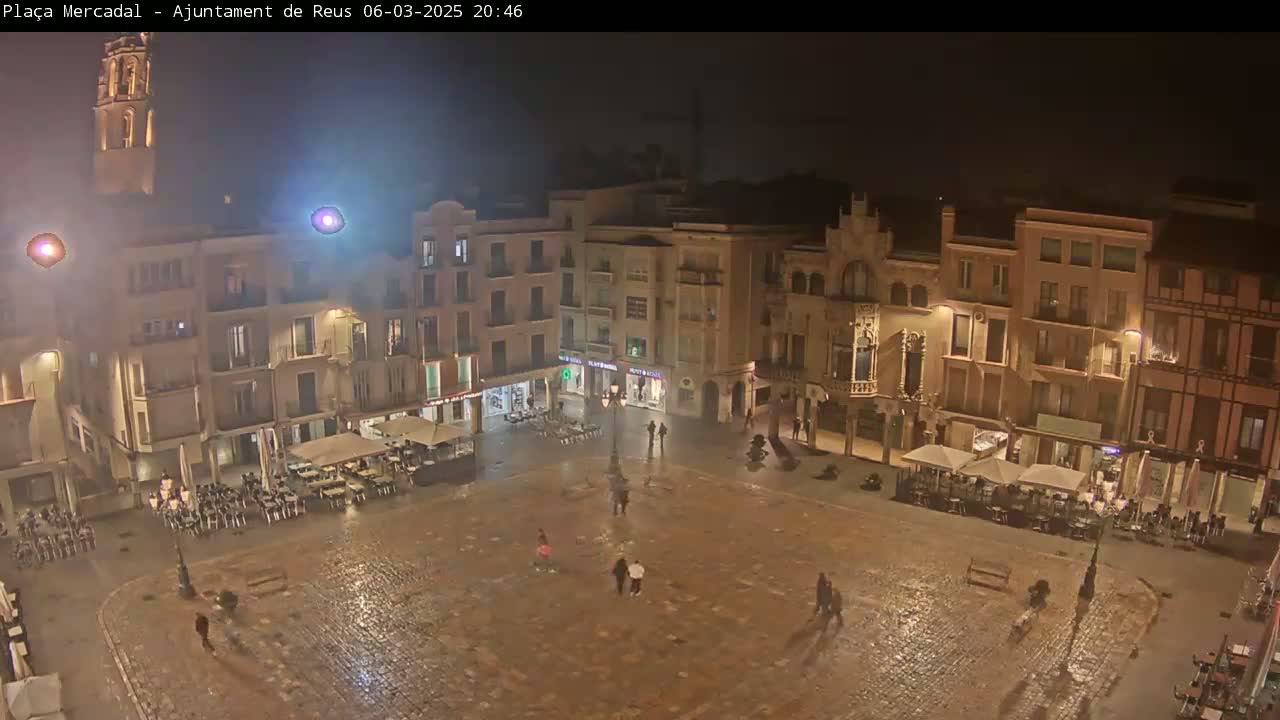 A wet, nighttime view of a town square with several buildings surrounding it, and people sitting at outdoor cafes.