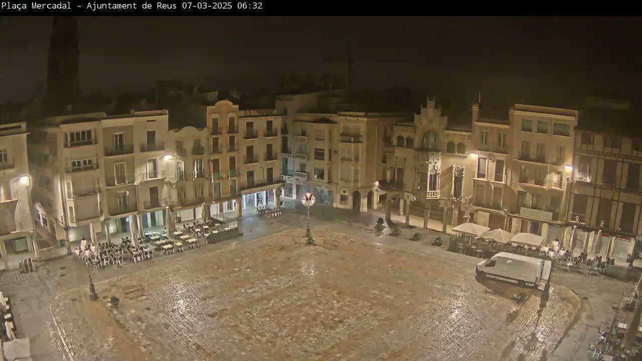 A wet, cobblestone town square at night is surrounded by multi-story buildings with outdoor seating areas.