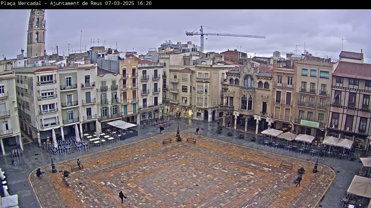 A large, paved town square with several buildings surrounding it, a few people walking around, and some outdoor cafe seating under umbrellas, on a cloudy day.