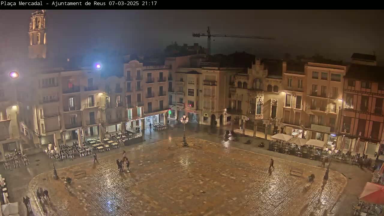 A rain-slicked town square at night, surrounded by illuminated buildings, with a few people and outdoor seating visible.