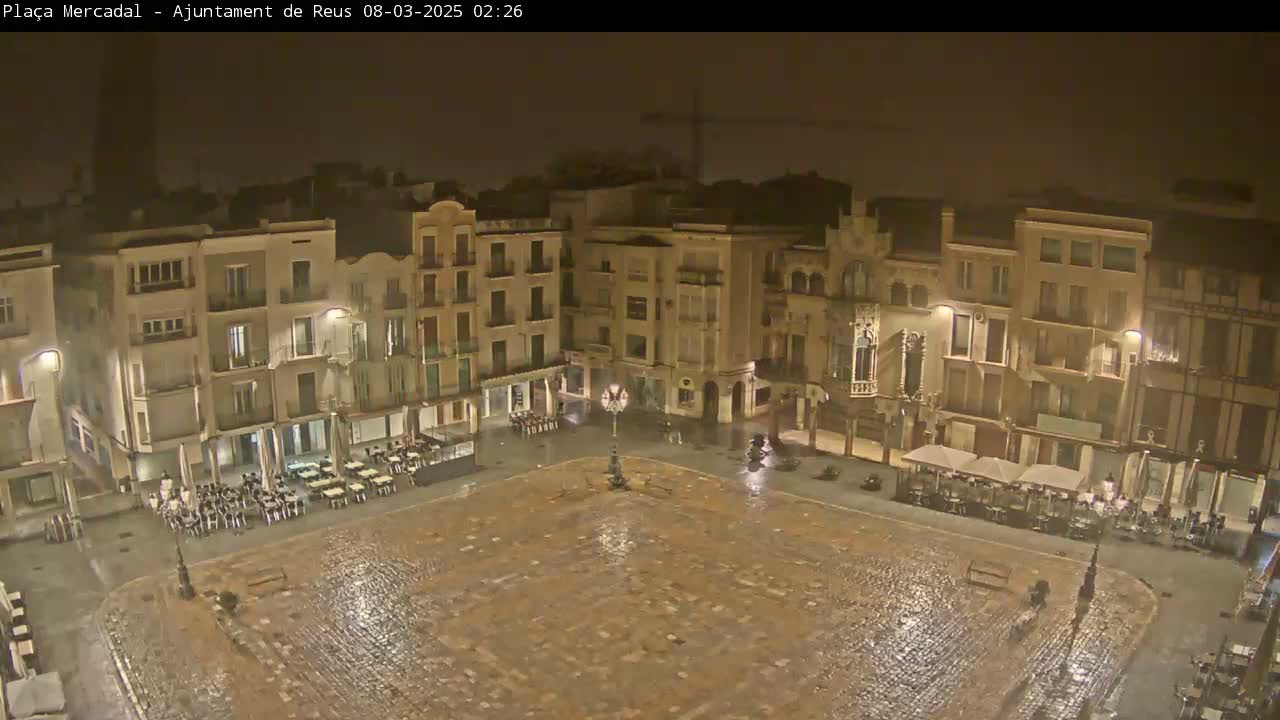 A rain-slicked town square at night, surrounded by multi-story buildings with outdoor seating areas.