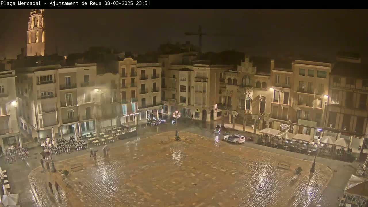 A rain-soaked town square at night is surrounded by illuminated buildings, with a few people and cars visible.