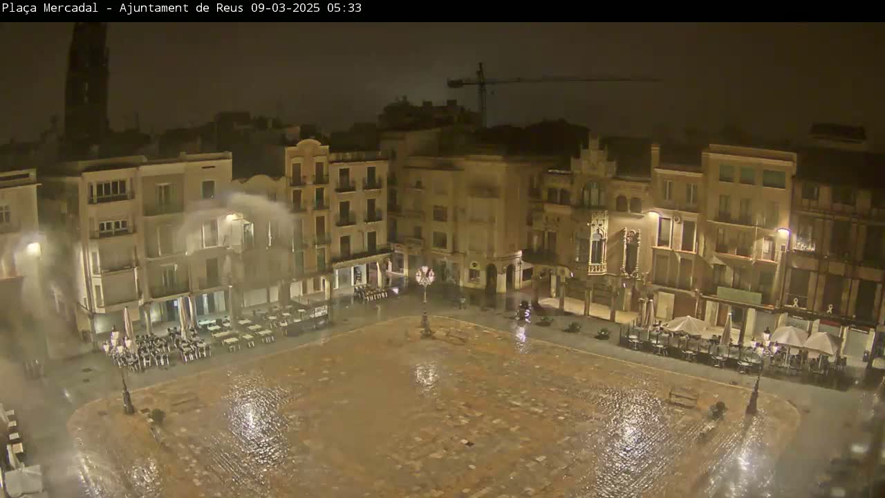 A nighttime view of a town square, wet from rain, with water spraying from fountains, and outdoor seating areas around the perimeter of the square.