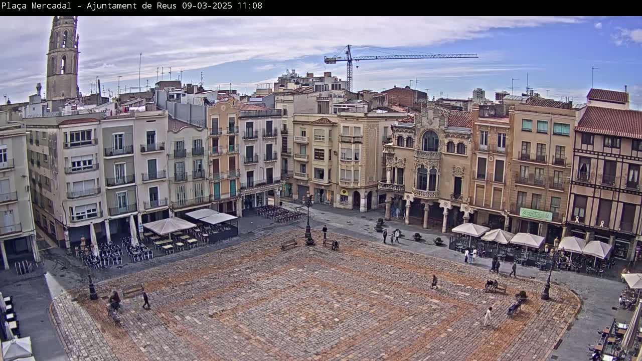 A large, cobblestone town square with several multi-story buildings surrounding it, under a partly cloudy sky.