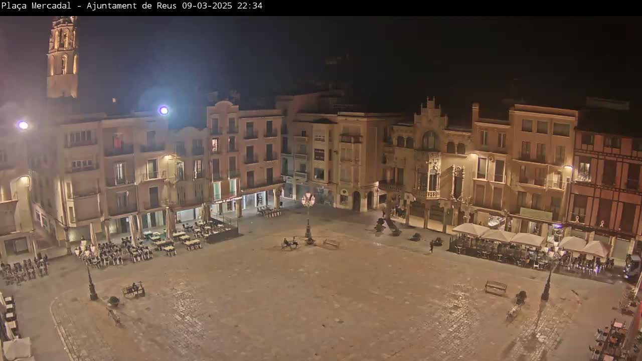 A mostly empty town square at night, surrounded by multi-story buildings with illuminated windows and outdoor seating areas.