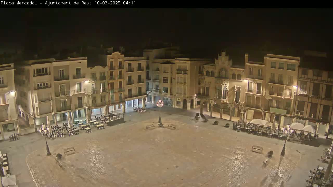 A nighttime view of a large, mostly empty town square surrounded by multi-story buildings with outdoor seating at some of the ground-floor businesses.