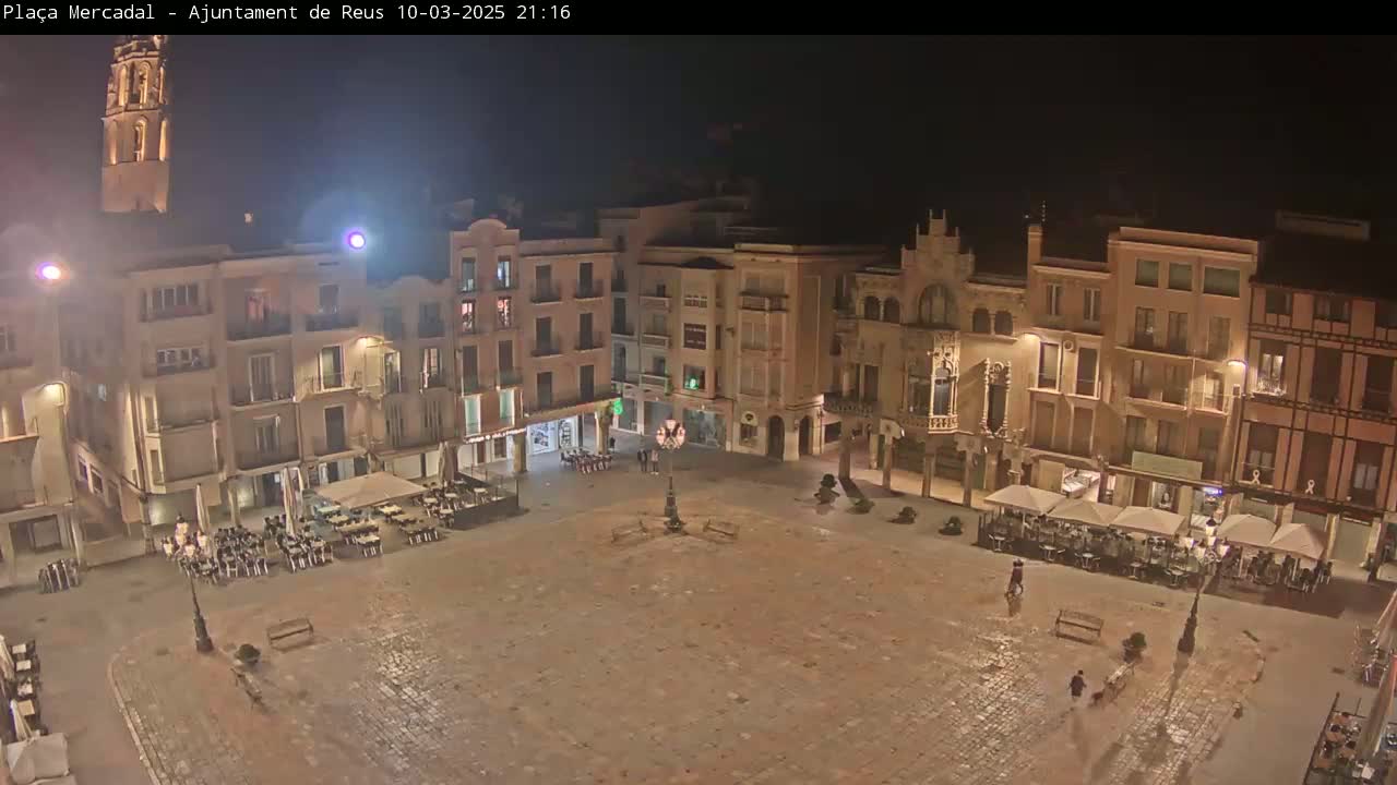 A nighttime view of a large, mostly empty town square surrounded by illuminated buildings, with a few people and outdoor seating areas visible.