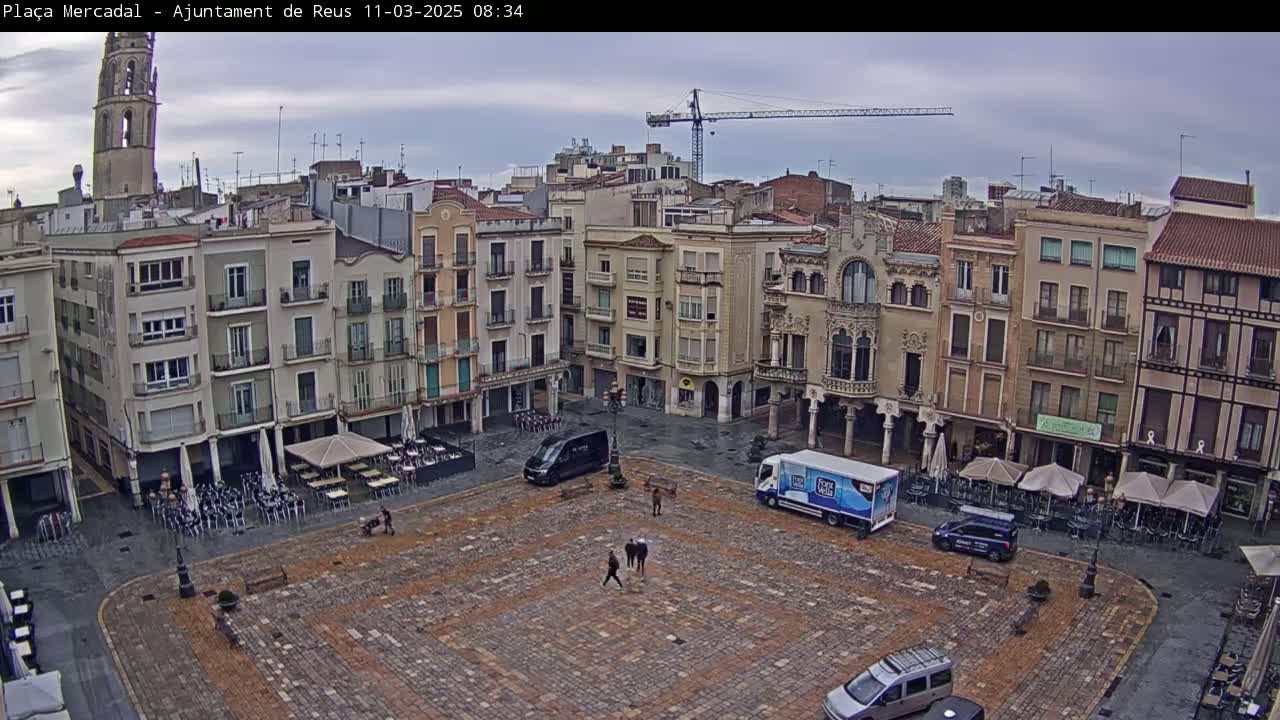 A paved town square, surrounded by multi-story buildings under an overcast sky, contains several parked vehicles, outdoor cafe seating, and a few pedestrians.