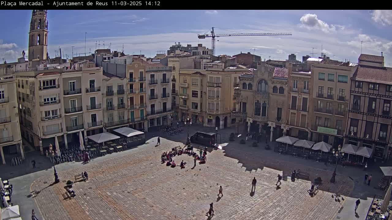 A sunny day reveals a large town square, surrounded by multi-story buildings, with people scattered throughout and outdoor seating areas under umbrellas.