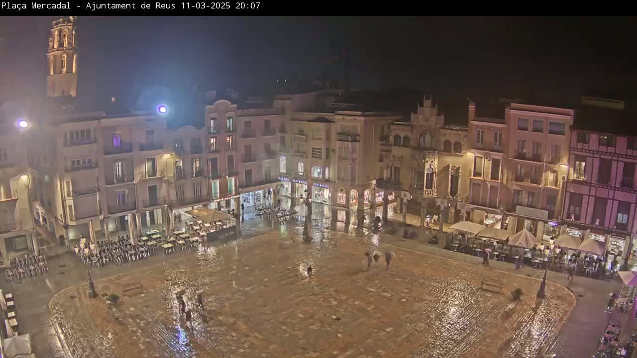 A rain-slicked town square at night, surrounded by illuminated buildings, with a few people walking and some outdoor seating areas.