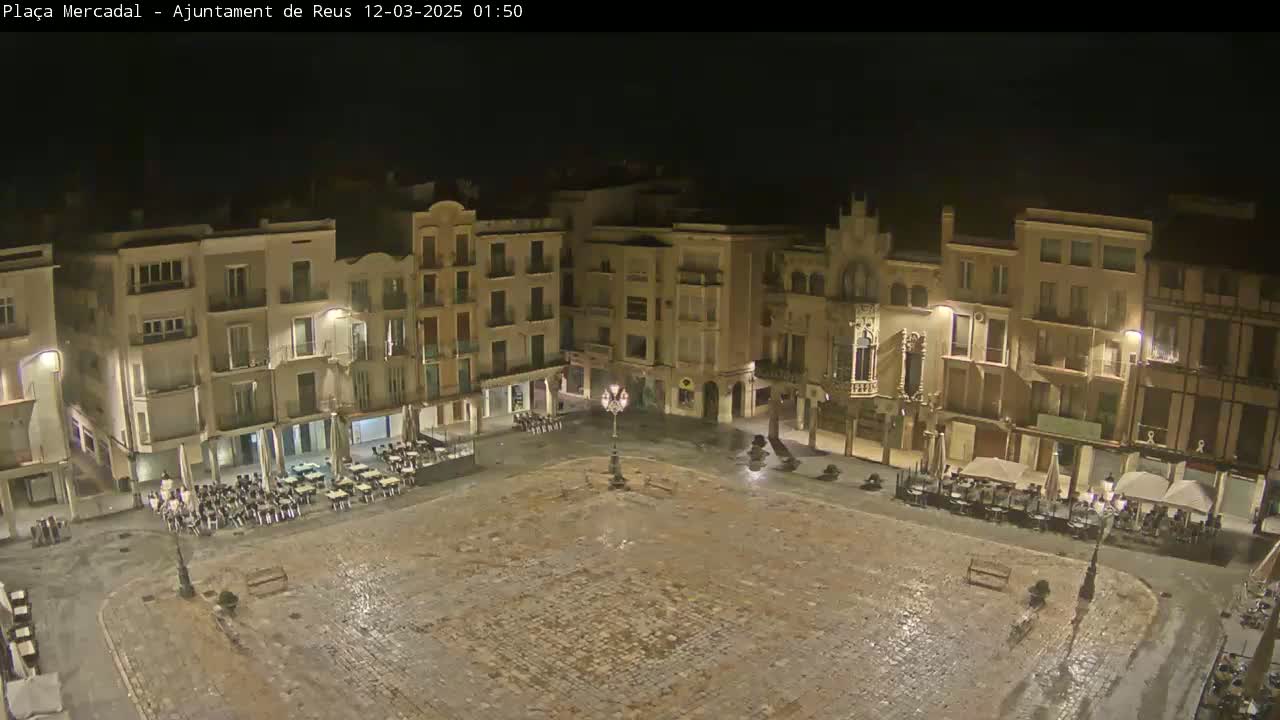 A nighttime, wet cobblestone town square is surrounded by multi-story buildings with outdoor seating areas at ground level.