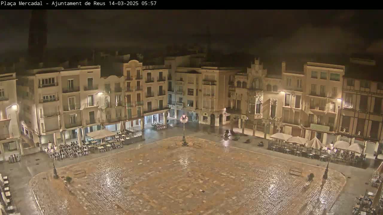 A rain-slicked, cobblestone town square at night, surrounded by multi-story buildings with outdoor seating areas under umbrellas.