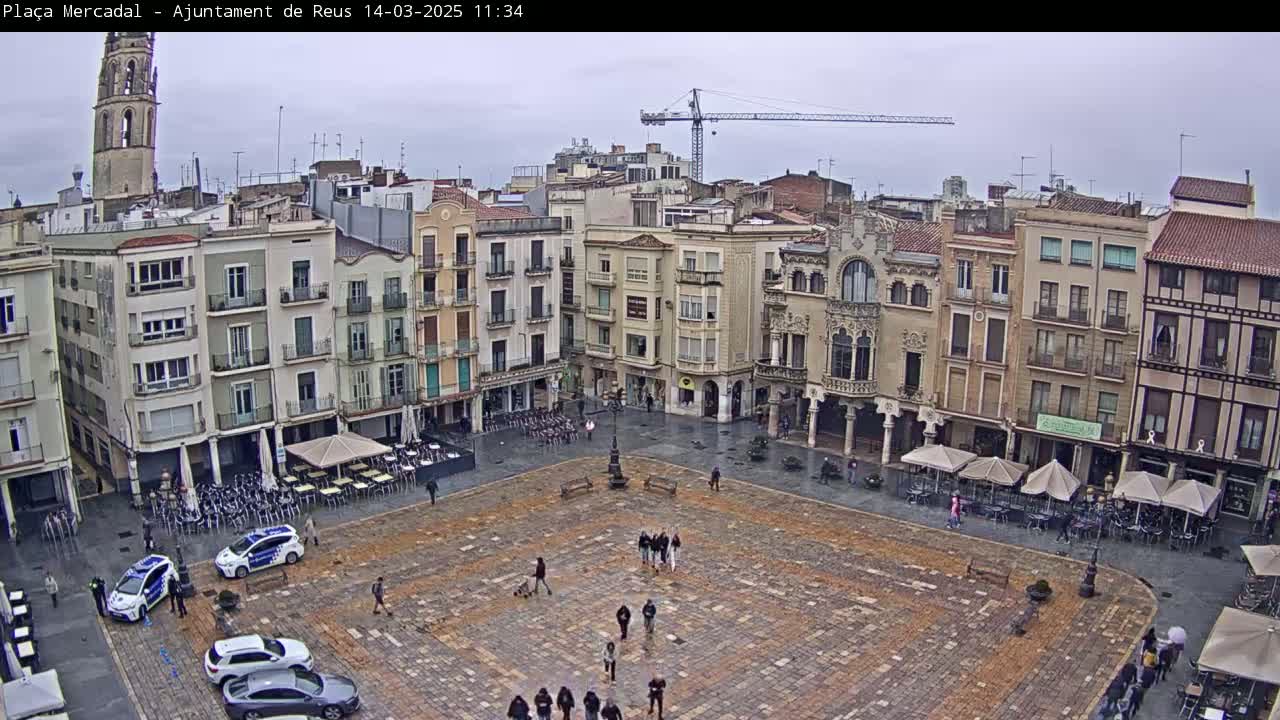 A large, paved town square with several buildings surrounding it, a few people walking around, and some parked police cars, under an overcast sky.