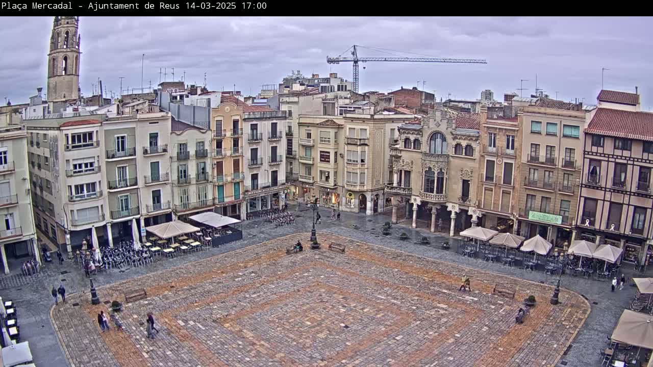 An overcast day reveals a large, cobblestone town square surrounded by multi-story buildings, some with outdoor seating areas, and a construction crane visible in the distance.