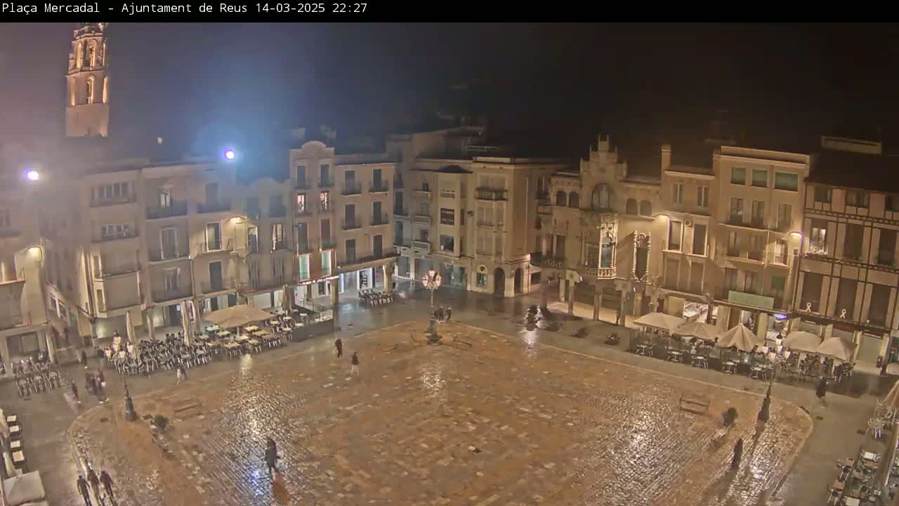 A rain-soaked town square at night, surrounded by illuminated buildings, with a few people sparsely scattered about.