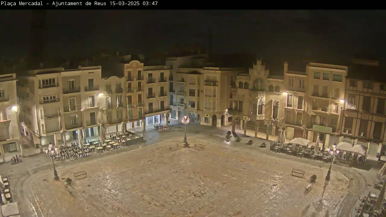 A large, empty town square at night, surrounded by multi-story buildings with some outdoor seating areas, is lightly dusted with snow or ice.
