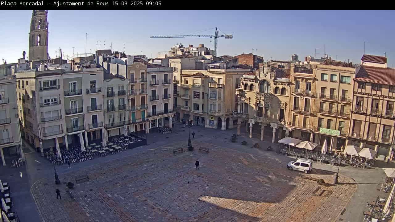 A large, mostly empty town square paved with stone, surrounded by multi-story buildings under a clear, sunny sky.