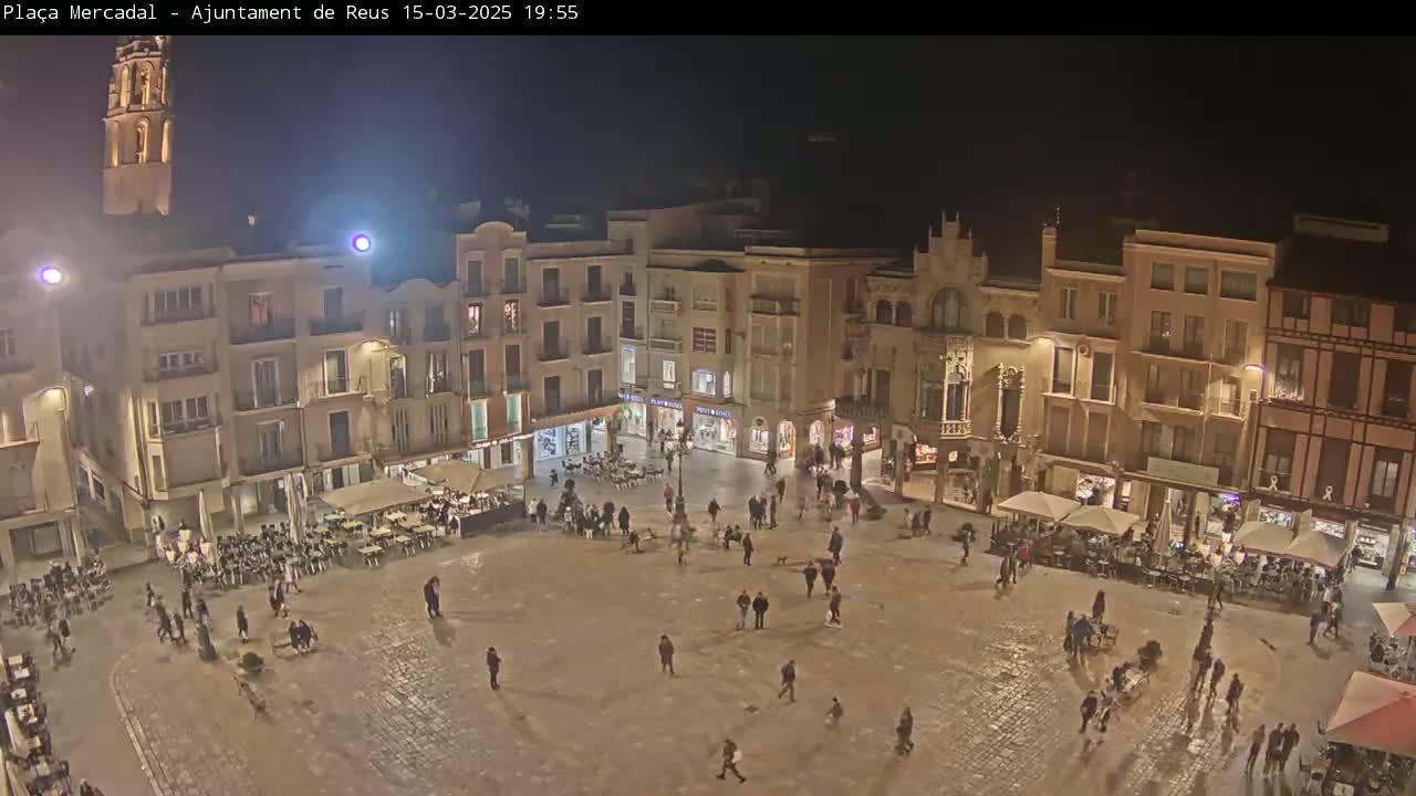 A nighttime, overhead view of a town square filled with people, illuminated buildings, and outdoor seating areas.