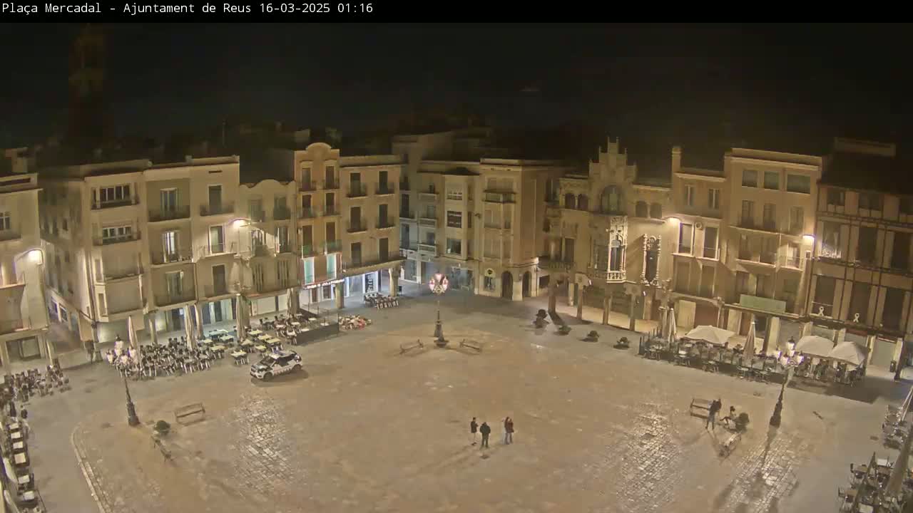 A mostly empty town square at night, illuminated by streetlights, is surrounded by multi-story buildings.