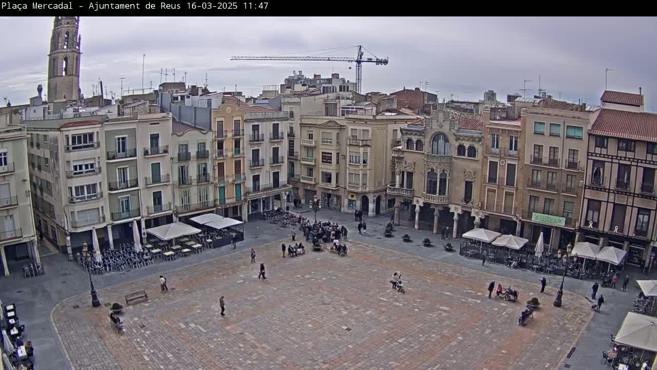 A large, paved town square is surrounded by multi-story buildings under an overcast sky, with a few people and outdoor seating at cafes visible.