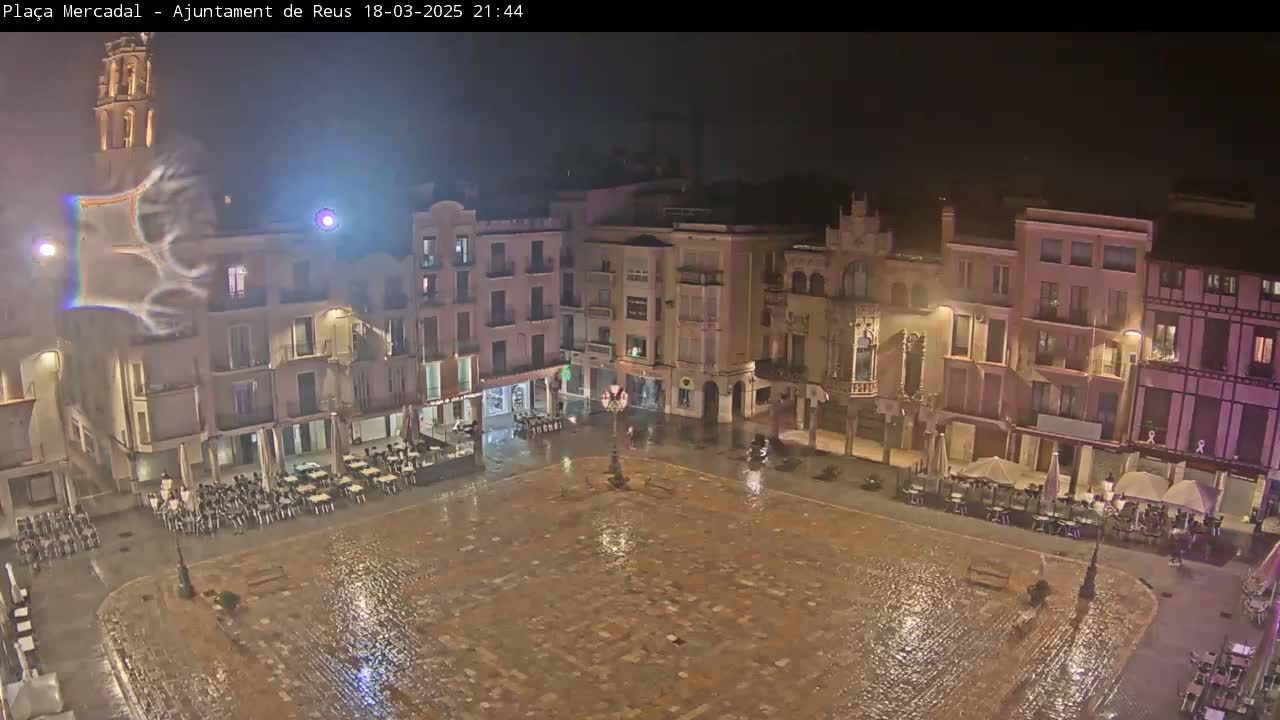 A rain-slicked town square at night, surrounded by multi-story buildings with outdoor seating areas.