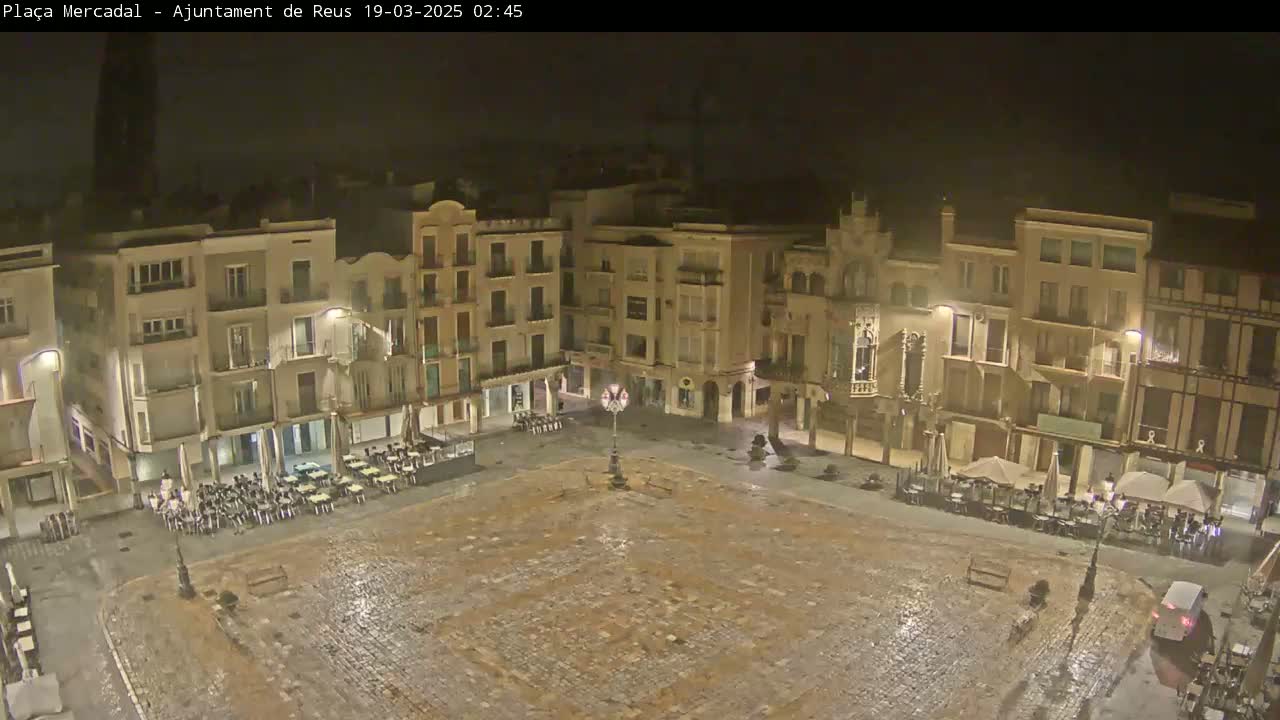 A rain-slicked, cobblestone town square at night, surrounded by multi-story buildings with outdoor seating areas.