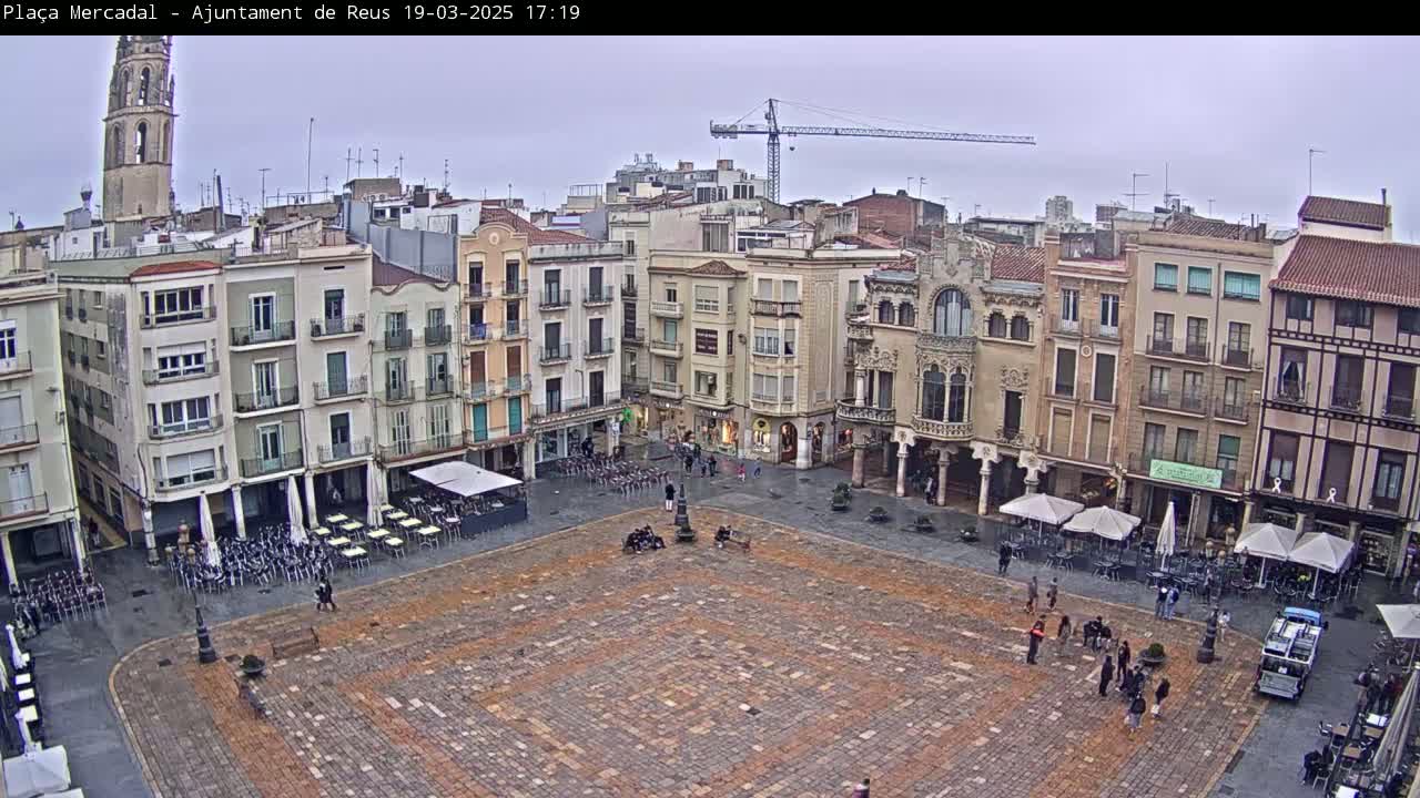 A large, paved town square is surrounded by multi-story buildings under an overcast sky, with a few people scattered throughout the square and outdoor seating areas at cafes.