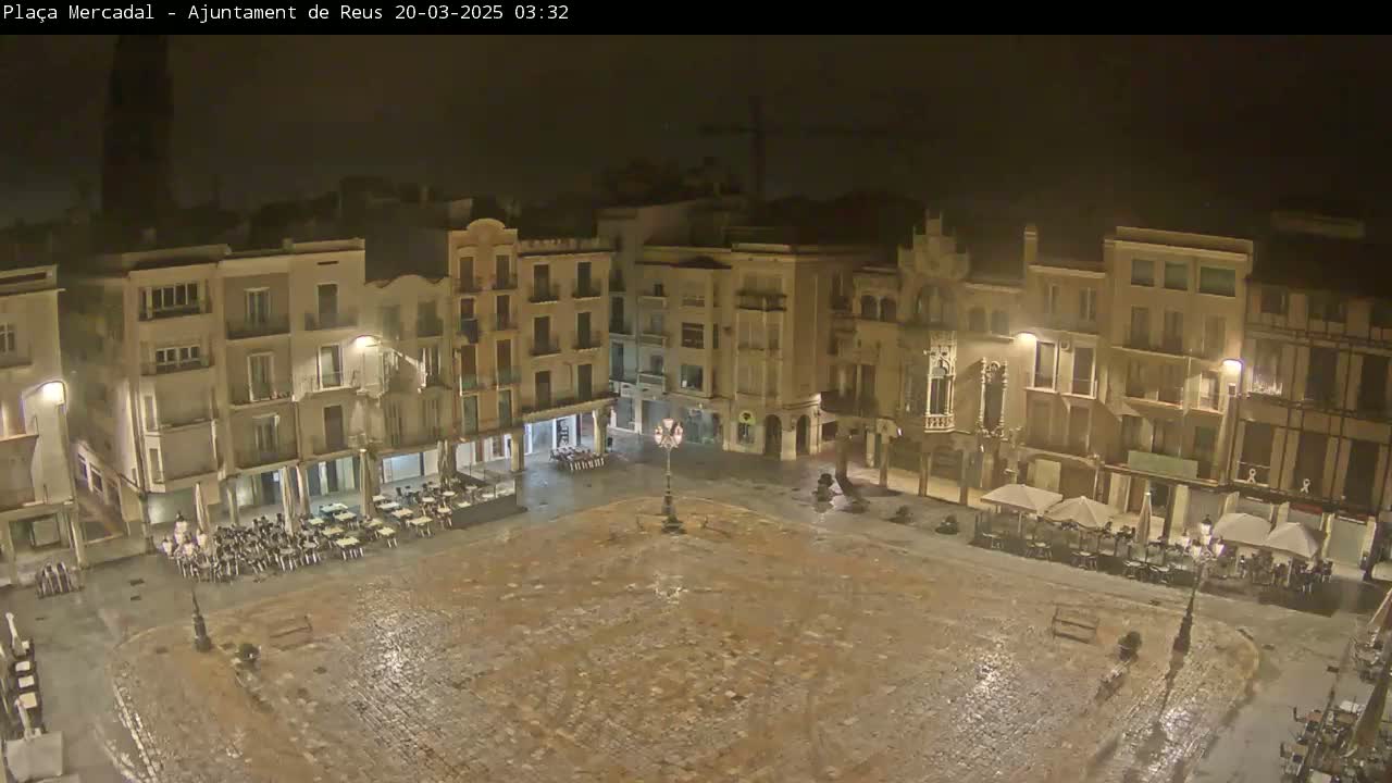 A nighttime view of a town square, wet from recent rain, with several buildings surrounding it and outdoor seating at various cafes.