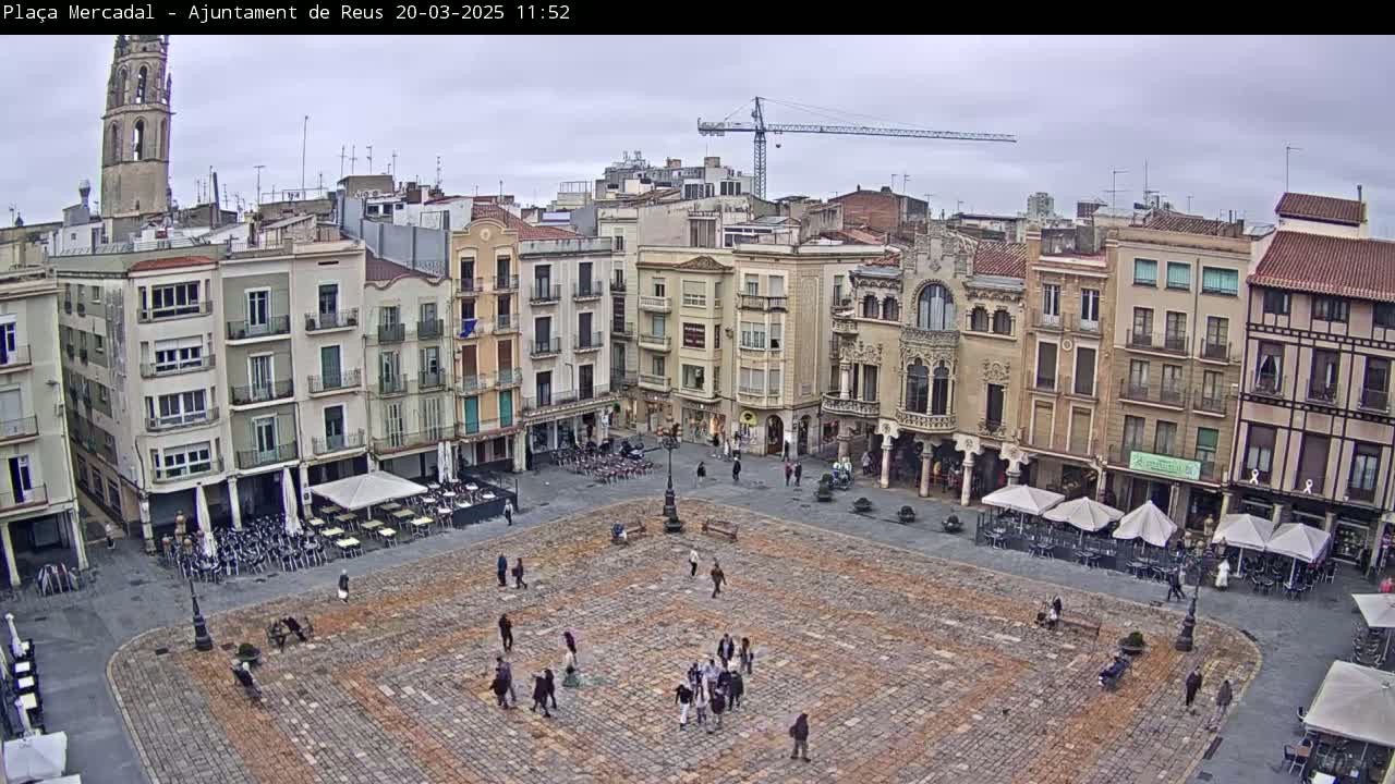 A large, cobblestone town square is surrounded by multi-story buildings under an overcast sky, with a few people walking around and outdoor seating at cafes.
