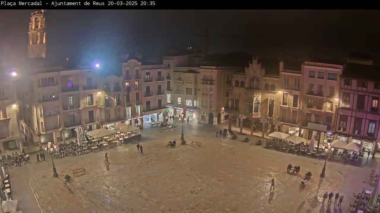 A nighttime view of a town square with several buildings surrounding a paved area, with some people and outdoor seating at cafes, under a dark, possibly rainy sky.