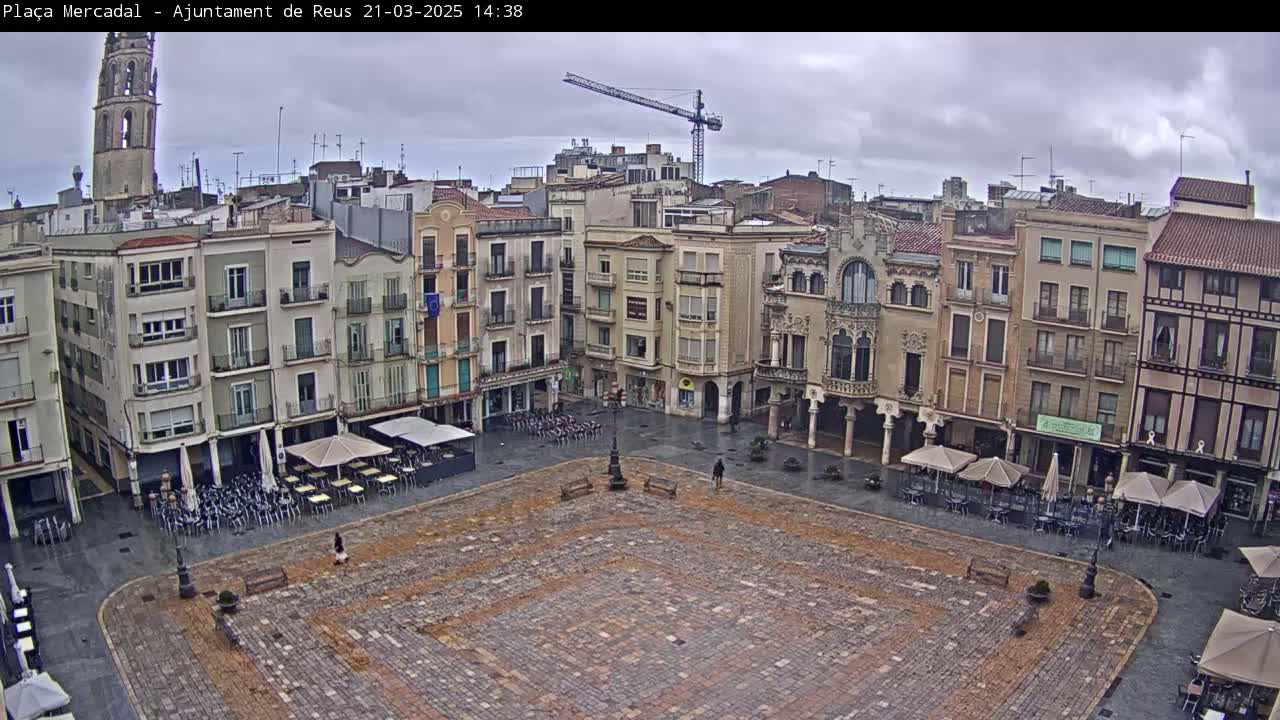 A large, paved town square is surrounded by multi-story buildings under an overcast sky, with some outdoor seating areas visible.