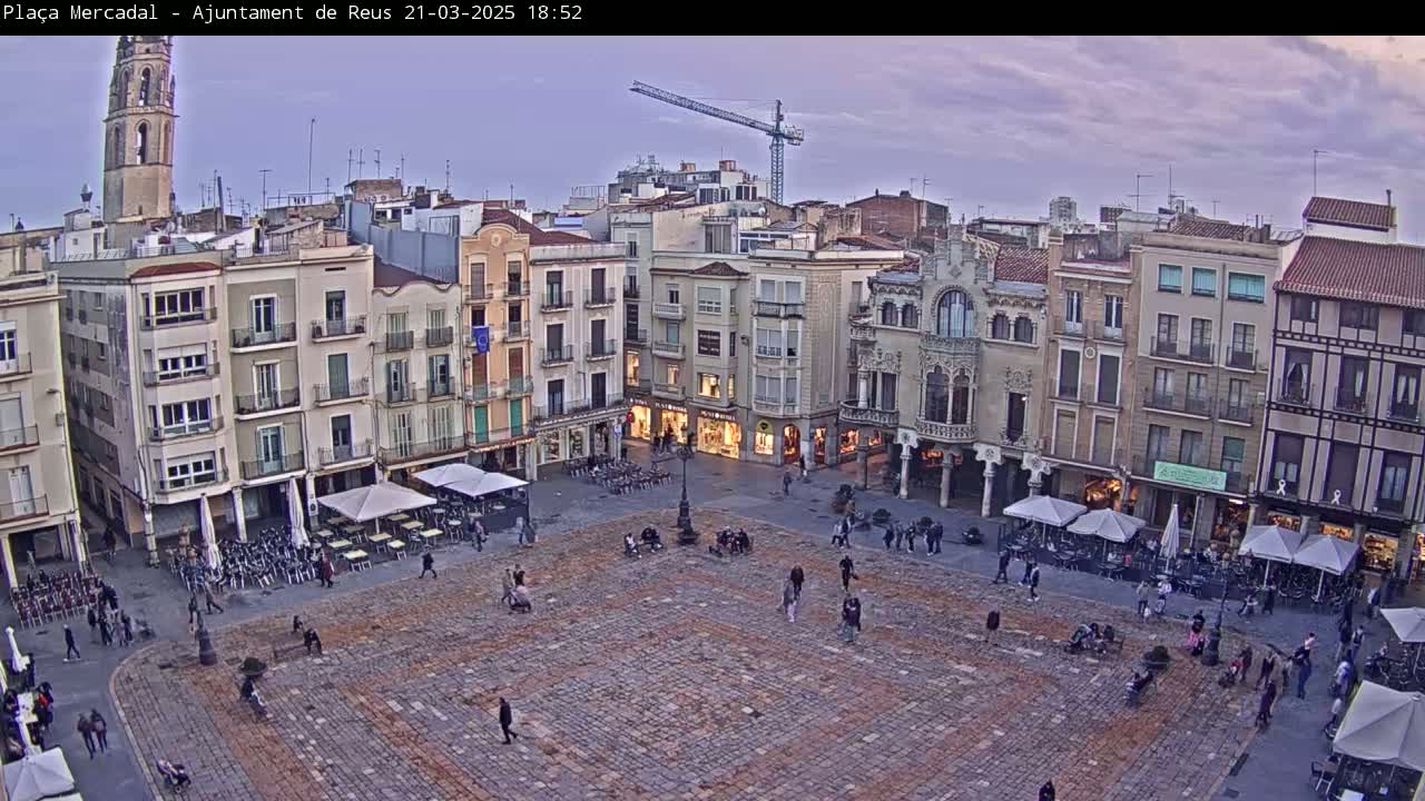 A large, cobblestone town square is populated with sparsely scattered people and several outdoor cafes under cloudy twilight skies, overlooked by multi-story buildings and a tall church tower.