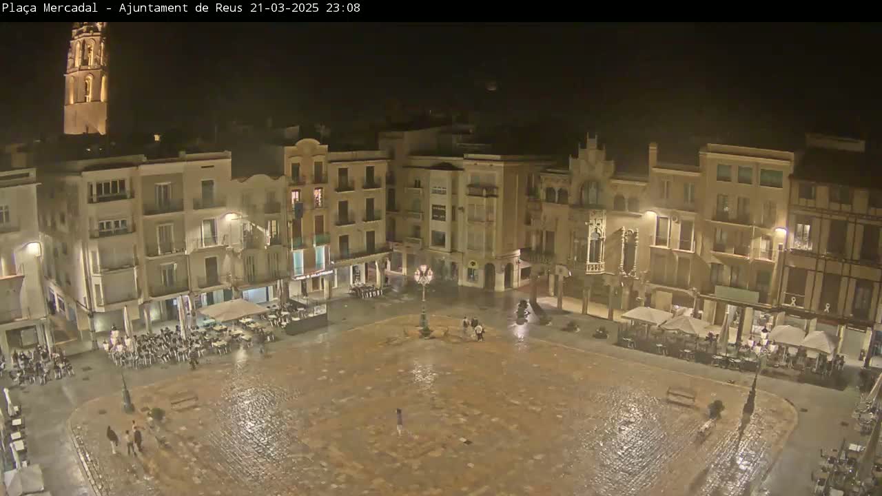 A nighttime view of a town square, illuminated by streetlights, showing several buildings surrounding a wet cobblestone plaza with a few people present.