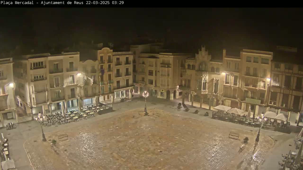 A nighttime, high-angle view of a large, empty cobblestone town square surrounded by multi-story buildings with outdoor seating areas at several of the establishments.