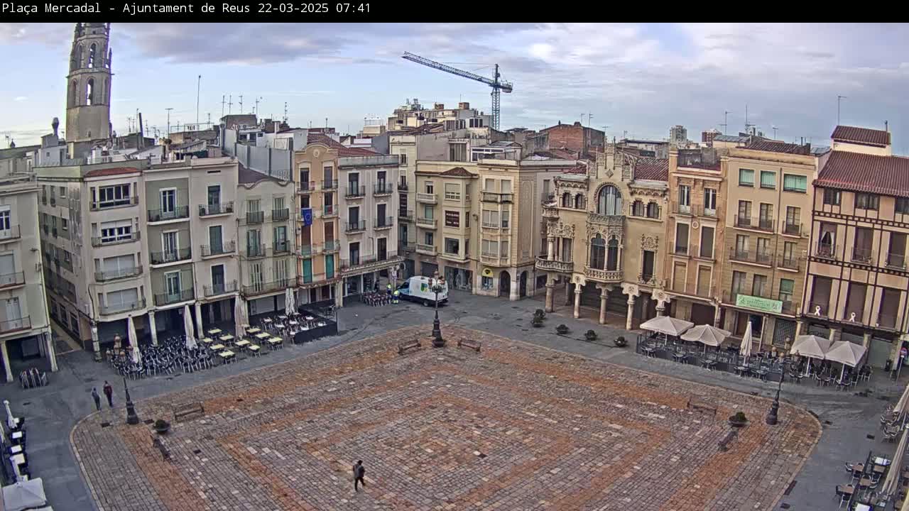A large, cobblestone town square with several multi-story buildings surrounding it under an overcast sky, with a construction crane visible in the background.