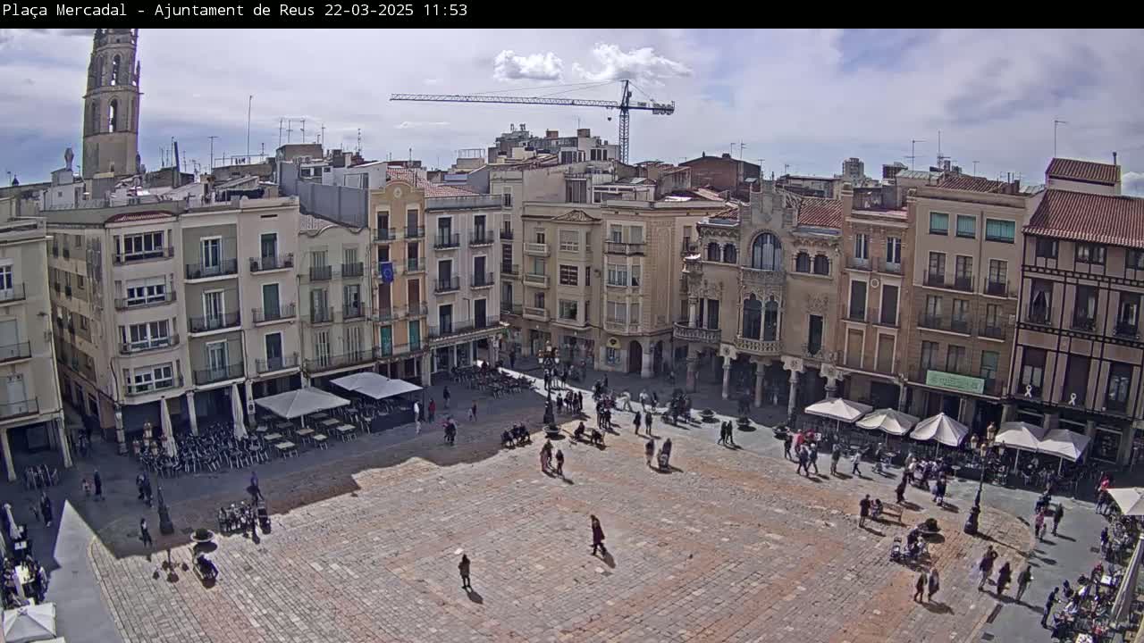 A partly cloudy day reveals a large, paved town square surrounded by multi-story buildings, with people scattered throughout and some outdoor seating areas.