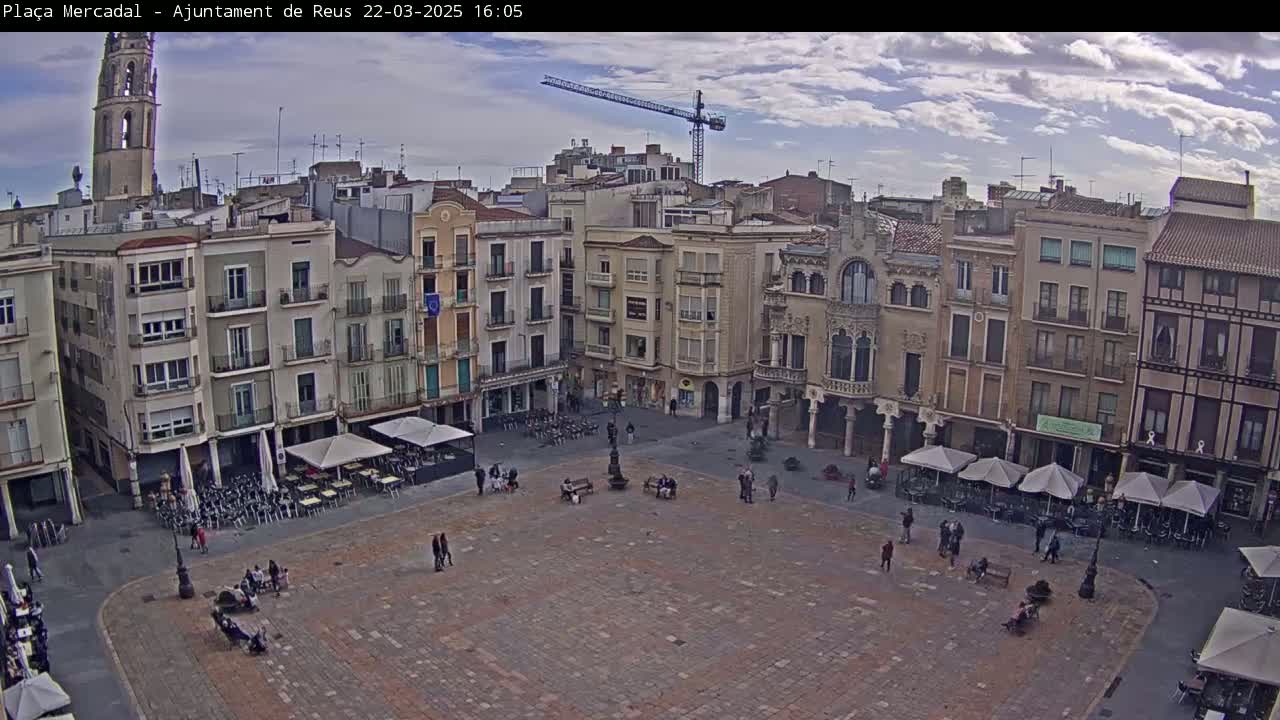 A large, mostly empty town square with surrounding buildings, outdoor seating areas, and a partly cloudy sky.