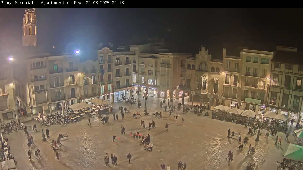 A nighttime view of a town square, filled with people and surrounded by illuminated buildings, under a clear, dark sky.