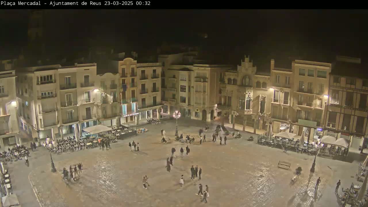 A nighttime view of a town square, surrounded by multi-story buildings, with several people scattered throughout.