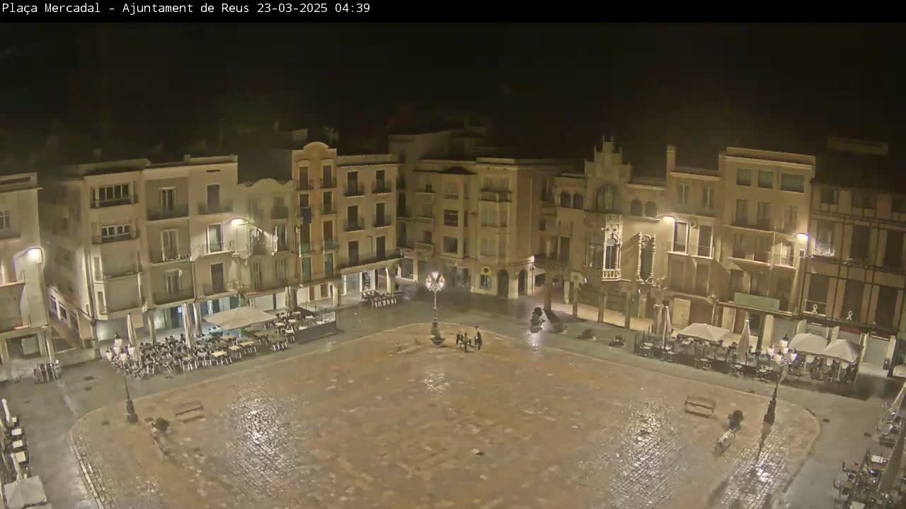 A wet, empty town square at night is surrounded by multi-story buildings with outdoor seating areas.