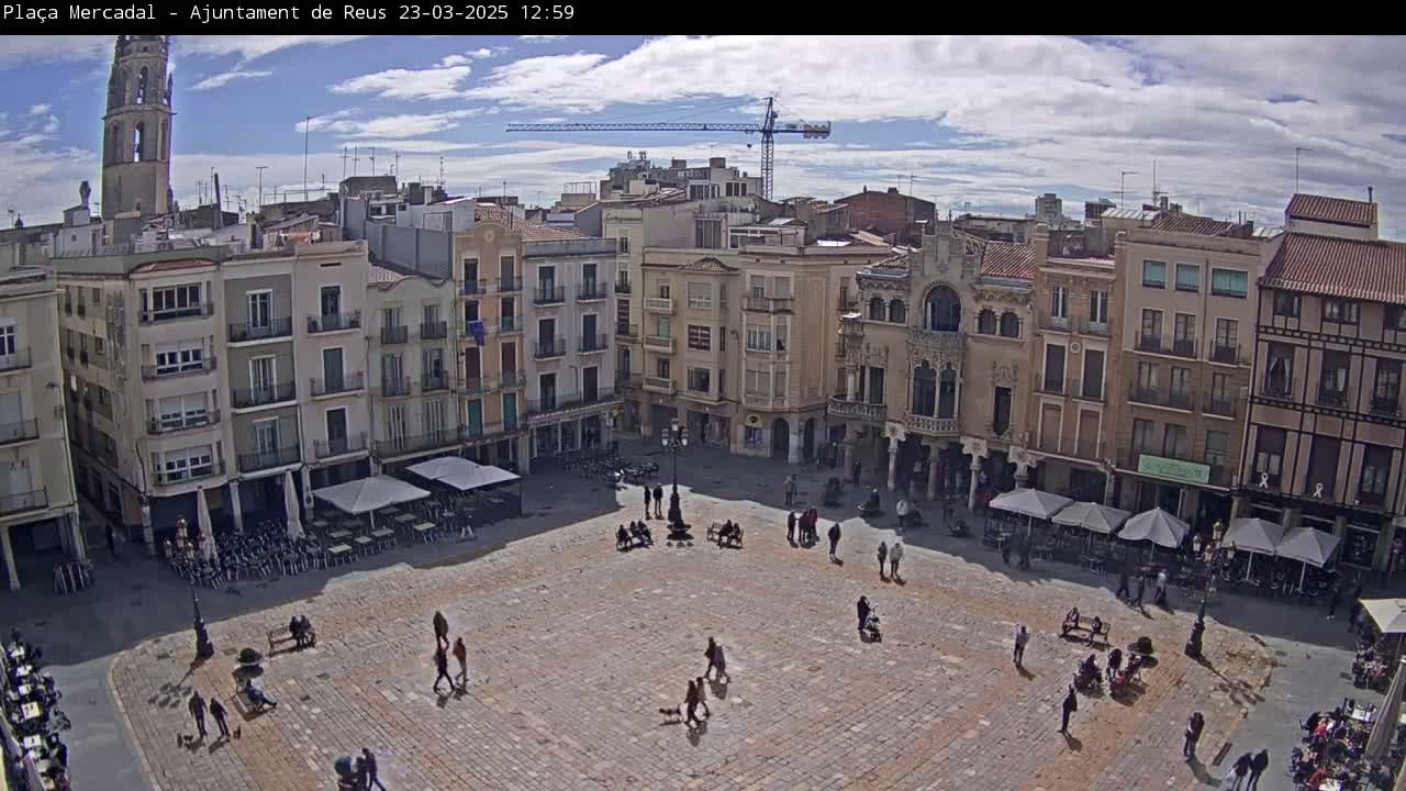 A mostly sunny day reveals a large town square, paved with brick, surrounded by multi-story buildings, with a few people walking around and several outdoor café umbrellas set up.
