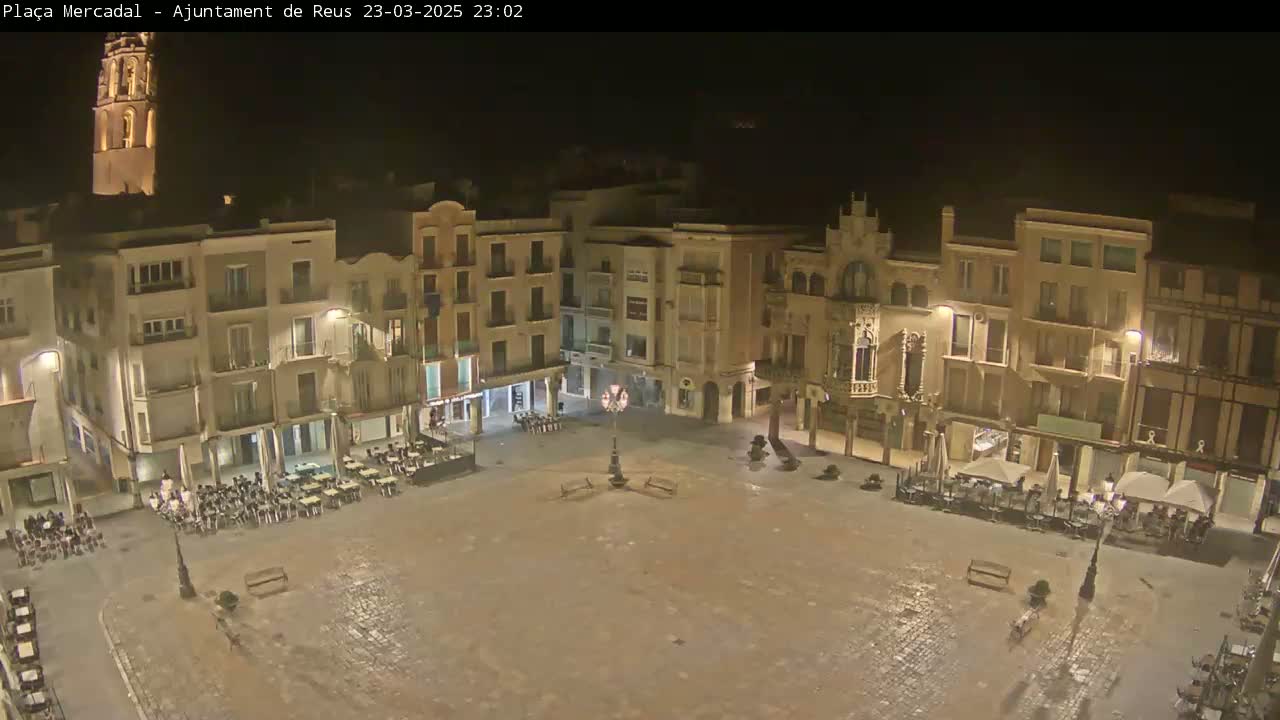 A mostly empty town square at night, surrounded by multi-story buildings, is illuminated under dark skies.