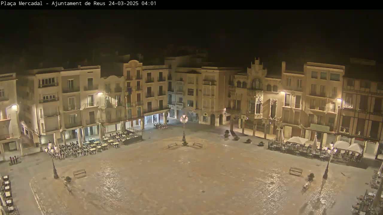 A mostly empty town square at night, illuminated by streetlights, is surrounded by multi-story buildings.