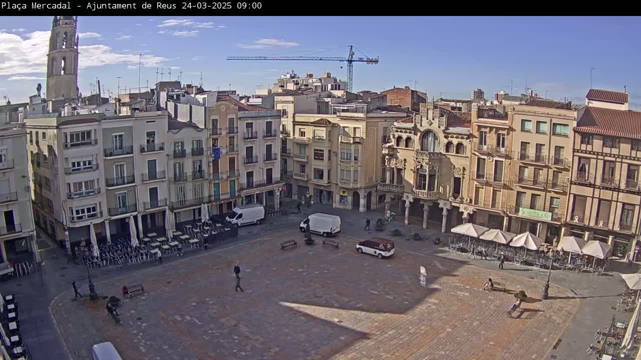 A mostly empty town square with several multi-story buildings surrounding it under a clear, sunny sky, and a construction crane visible in the background.