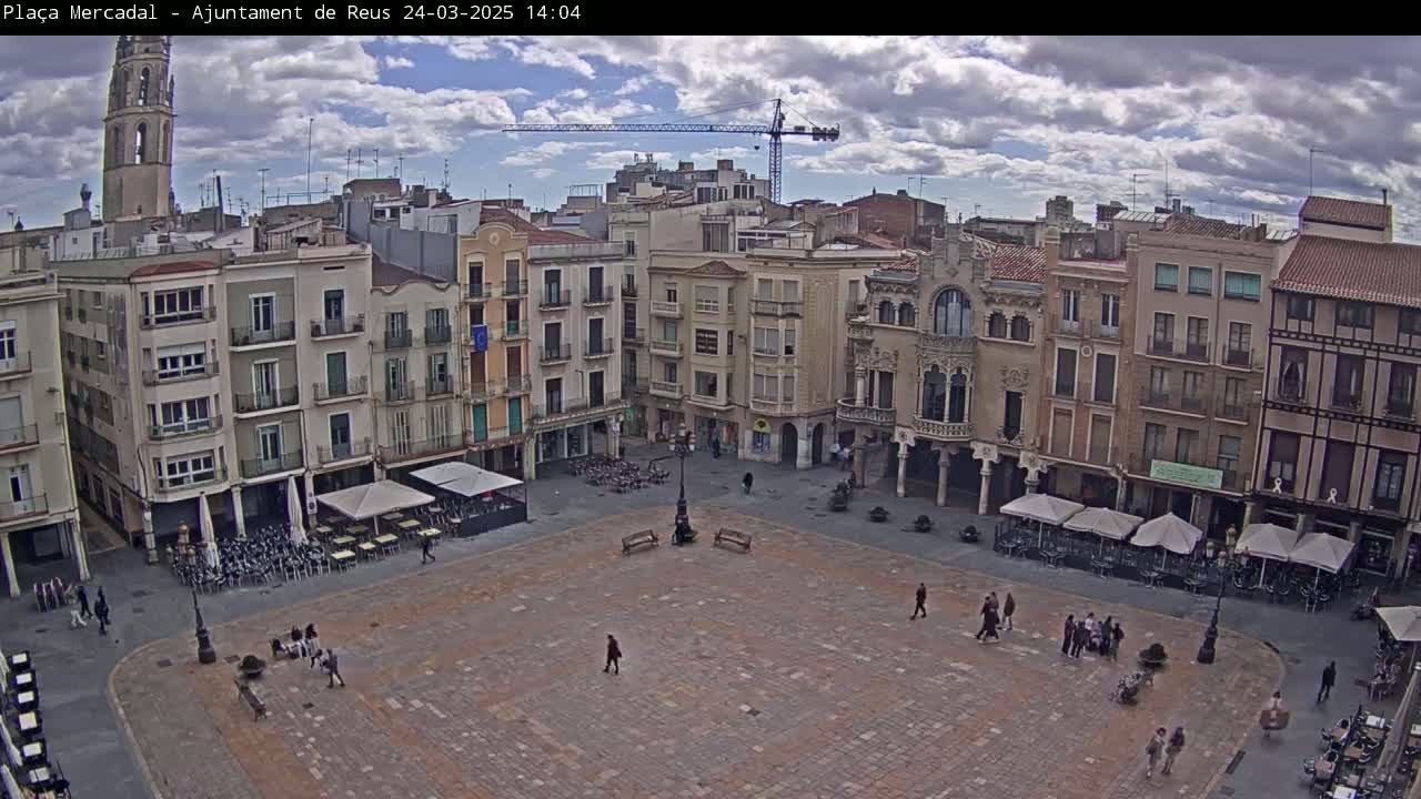 A mostly empty town square with surrounding buildings, outdoor cafes, and a construction crane visible in the distance under a partly cloudy sky.