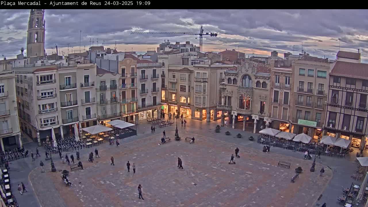 A large town square with several buildings surrounding it, under a cloudy evening sky, is sparsely populated with people.