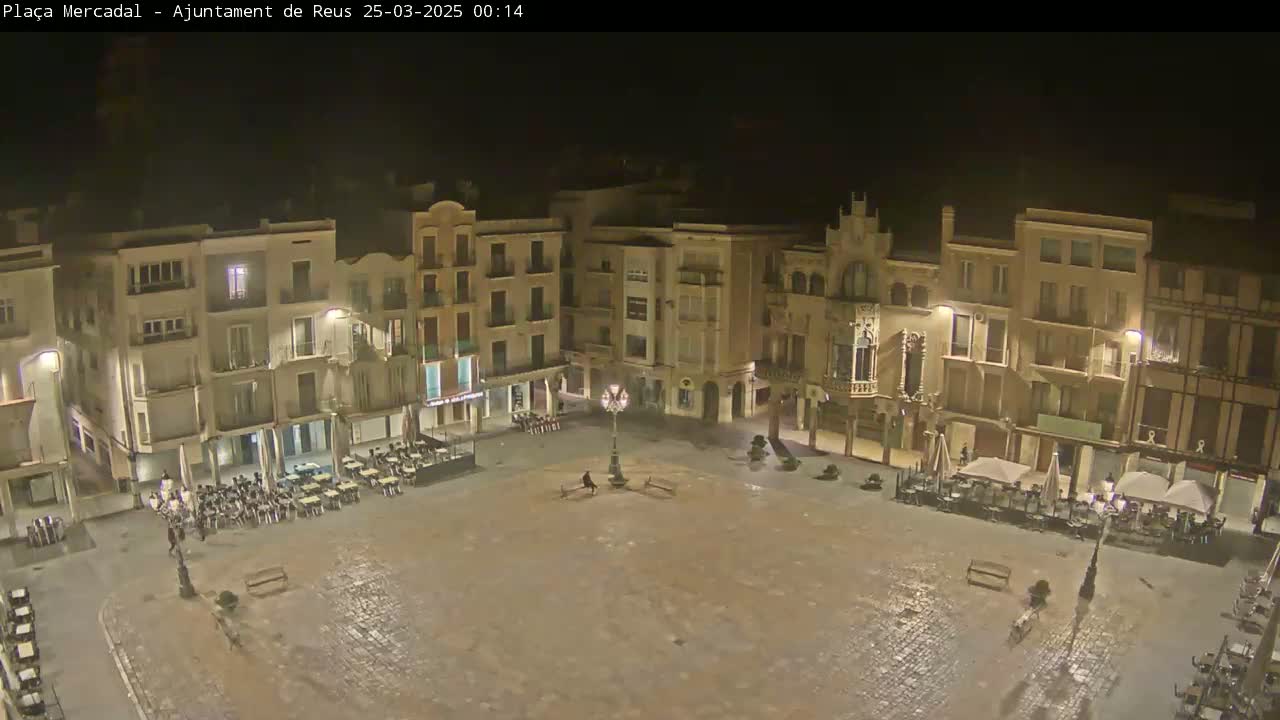 A nighttime, empty town square is surrounded by multi-story buildings with outdoor seating areas in front of some of them.