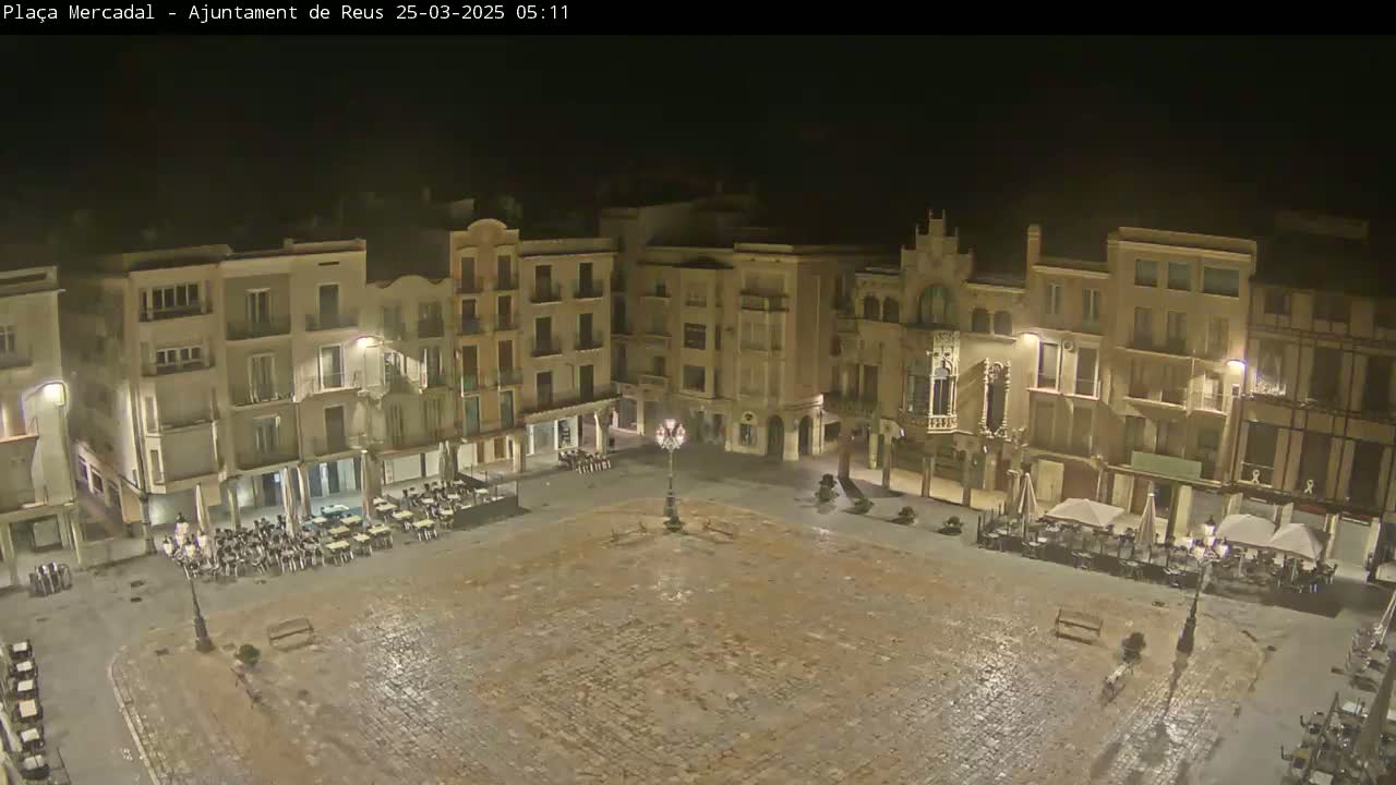 A large, empty cobblestone town square at night, surrounded by multi-story buildings with outdoor seating areas, is lightly wet.